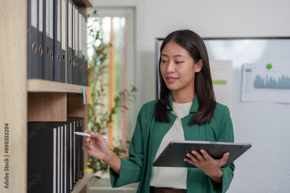 Attractive Asian woman standing and pulling a file Look through the files on the shelf to find financial documents in the meeting. Presentation of work within the company, management concepts. Attractive Asian woman standing and pulling a file Look through the files on the shelf to find financial documents in the meeting. Presentation of work within the company, management concepts.
