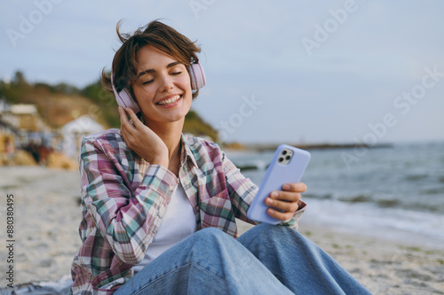 Young calm woman wear shirt casual clothes headphones sits listen music use mobile cell phone listen music rest on sea ocean sand shore beach outdoor seaside in summer day free time Lifestyle concept #880380495