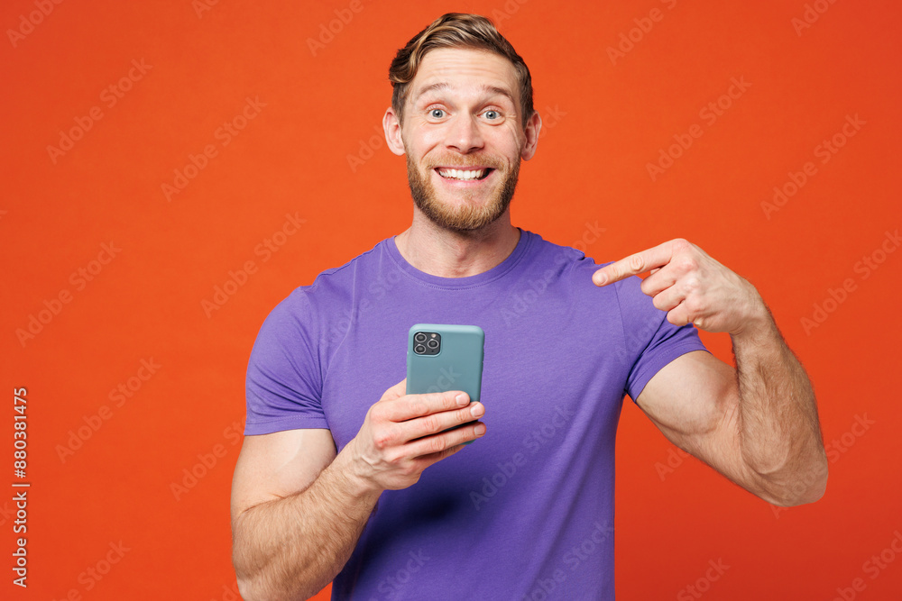 Young surprised shocked happy man he wear purple t-shirt casual clothes hold in hand use point index finger on mobile cell phone isolated on red orange background studio portrait. Lifestyle concept Young surprised shocked happy man he wear purple t-shirt casual clothes hold in hand use point index finger on mobile cell phone isolated on red orange background studio portrait. Lifestyle concept