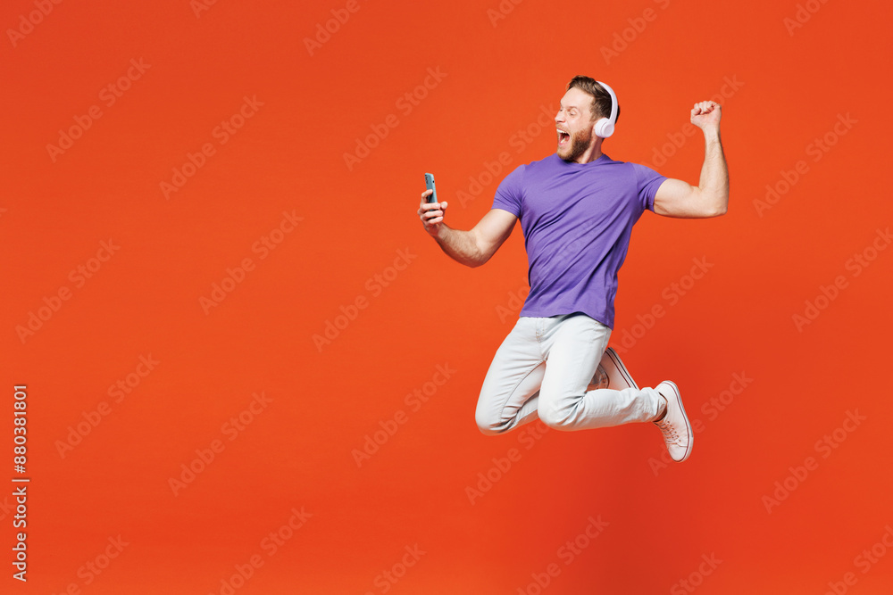 Full body young man wear purple t-shirt casual clothes jump high hold in hand use mobile cell phone listen to music in headphones do winner gesture isolated on red orange background Lifestyle concept Full body young man wear purple t-shirt casual clothes jump high hold in hand use mobile cell phone listen to music in headphones do winner gesture isolated on red orange background Lifestyle concept