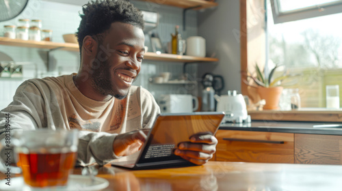 An African black man is sitting at a table with a laptop and a cup of tea. He looks through the morning news and mail on his tablet