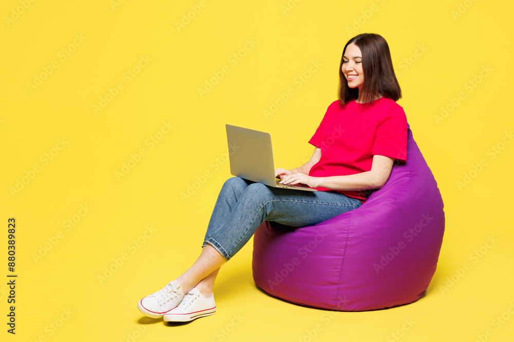 Full body young smart IT woman wear pink t-shirt casual clothes sit in bag chair hold use work on laptop pc computer chat online isolated on plain yellow orange background studio. Lifestyle concept. Full body young smart IT woman wear pink t-shirt casual clothes sit in bag chair hold use work on laptop pc computer chat online isolated on plain yellow orange background studio. Lifestyle concept.