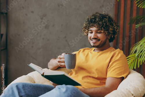 Young smiling fun Indian man wear orange casual clothes reading novel book drink coffee sits in armchair stay at home hotel flat rest relax spend free spare time in living room indoor. Lounge concept.