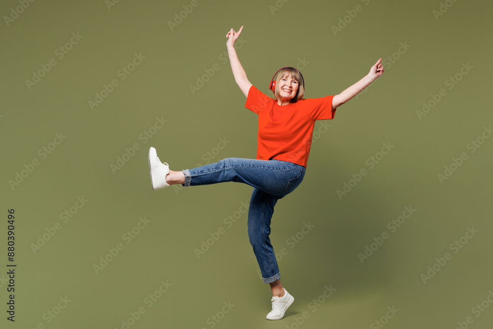 Full body elderly blonde woman 50s years old she wears orange t-shirt casual clothes listen to music in headphones raise up hands isolated on plain pastel green background studio. Lifestyle concept. Full body elderly blonde woman 50s years old she wears orange t-shirt casual clothes listen to music in headphones raise up hands isolated on plain pastel green background studio. Lifestyle concept.