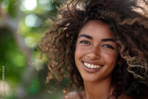 A smiling, multiethnic woman with curly hair blowing in the wind. The background is blurred with green foliage