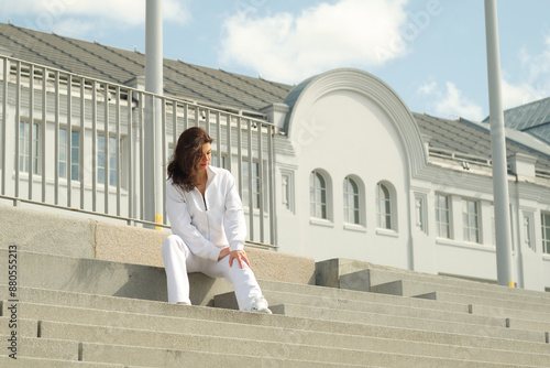 emale in a white tracksuit, seated on concrete steps with a modern building in the background. Concept: Demonstrating how urban environments can provide spaces for relaxation and reflection, promoting photo