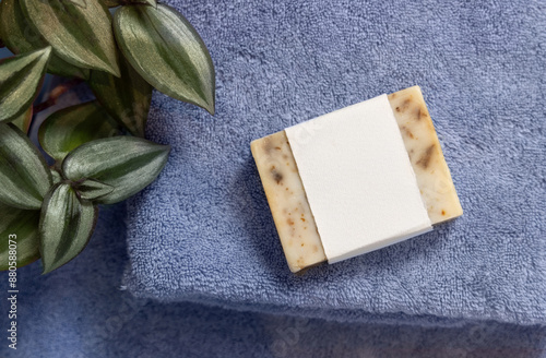 Soap bar with blank label on blue folded towel near green plant top view, packaging mockup photo