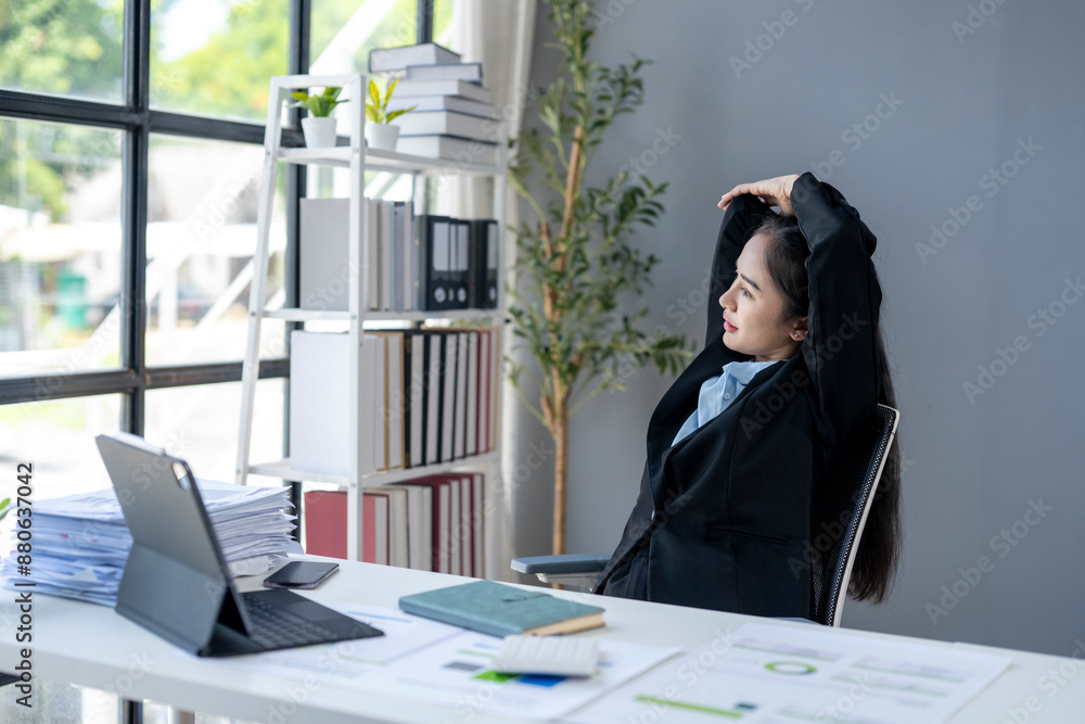 A woman in a black suit is sitting at a desk with a laptop and a stack of papers A woman in a black suit is sitting at a desk with a laptop and a stack of papers