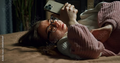 Bored teen girl in glasses using smartphone, wait for important message, interesting information update. Relationship problem. Move phone away with upset. Lying on bed in a sad bad mood