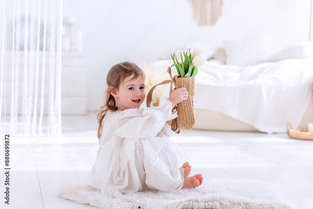 Little girl sits on floor in white Boho minimalist interior. Toddler in dress, nightie holds bouquet of spring tulips in hands, hugs, smells flowers. Airy, light filled space. Pampas grass decor Little girl sits on floor in white Boho minimalist interior. Toddler in dress, nightie holds bouquet of spring tulips in hands, hugs, smells flowers. Airy, light filled space. Pampas grass decor