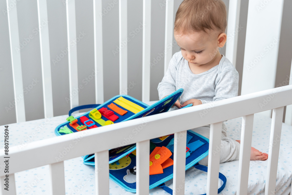 Baby playing with montessori busy book sitting in crib. Concept of smart books and modern toys Baby playing with montessori busy book sitting in crib. Concept of smart books and modern toys