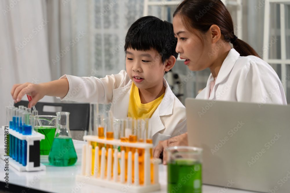 Boy in white lab coat and yellow shirt conducting science experiment and handling test tubes while sitting with Asian woman teacher or mentor at laboratory room Boy in white lab coat and yellow shirt conducting science experiment and handling test tubes while sitting with Asian woman teacher or mentor at laboratory room