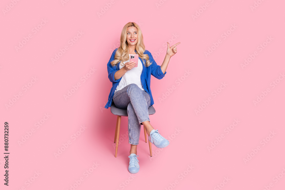 Full size photo of cheerful woman dressed shirt sit on chair hold smartphone directing look empty space isolated on pink color background Full size photo of cheerful woman dressed shirt sit on chair hold smartphone directing look empty space isolated on pink color background