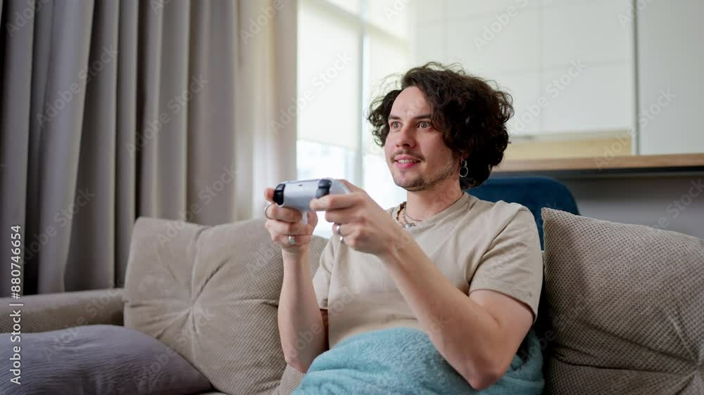 Happy brunette guy with curly hair and mustache plays video games and wins with a joystick and sits on the sofa at home in a modern apartment Happy brunette guy with curly hair and mustache plays video games and wins with a joystick and sits on the sofa at home in a modern apartment