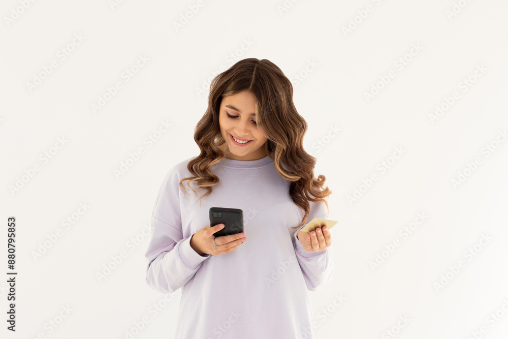 Cheerful young girl showing plastic credit card while using mobile phone isolated over white background, wearing purple sweatshirt Cheerful young girl showing plastic credit card while using mobile phone isolated over white background, wearing purple sweatshirt