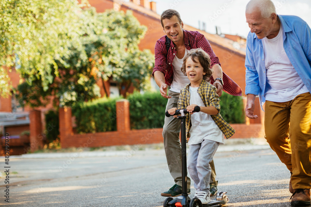 Fototapeta premium Father and grandfather teaching young boy to ride a scooter on suburban street
