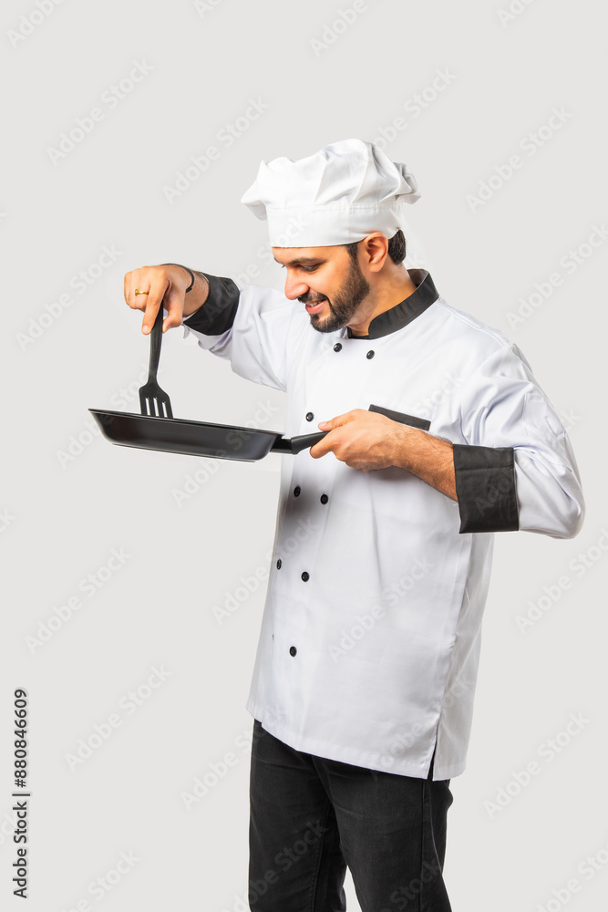 Indian asian male Chef in uniform happily holding spatula and frying pan on white background Indian asian male Chef in uniform happily holding spatula and frying pan on white background