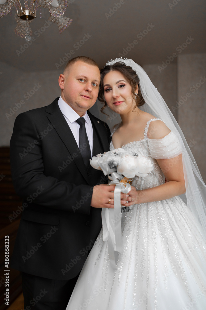 A bride and groom are posing for a picture in front of a chandelier. The bride is wearing a white dress and the groom is wearing a black suit A bride and groom are posing for a picture in front of a chandelier. The bride is wearing a white dress and the groom is wearing a black suit