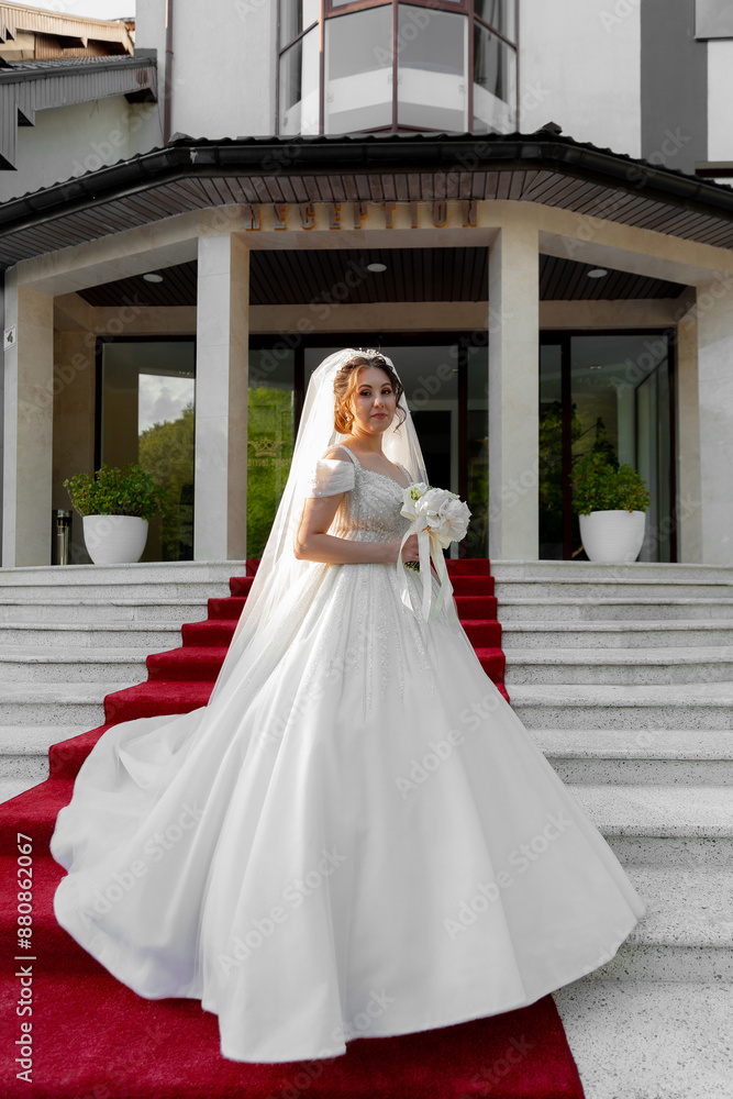 A bride is walking down a red carpet in a white dress. She is holding a bouquet of flowers and a white veil. The bride is wearing a long white gown with a long train A bride is walking down a red carpet in a white dress. She is holding a bouquet of flowers and a white veil. The bride is wearing a long white gown with a long train