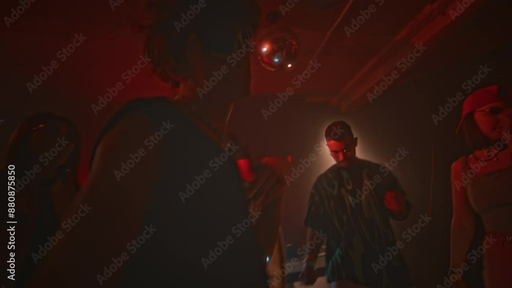 Handheld lowkey shot of young charismatic guy in mesh tank top and techno rave sunglasses making energetic powerful movements to camera while dancing to EDM among friends in red neon lit nightclub Handheld lowkey shot of young charismatic guy in mesh tank top and techno rave sunglasses making energetic powerful movements to camera while dancing to EDM among friends in red neon lit nightclub