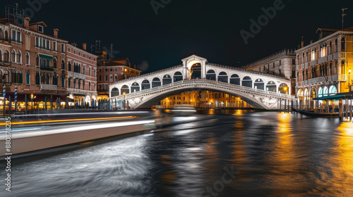 A serene nighttime view of Venice's iconic Rialto Bridge, beautifully illuminated, with blurred lights of passing boats reflecting on the calm Grand Canal. © VK Studio