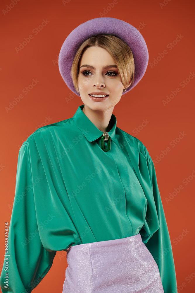 A stylish woman poses in a green shirt and lavender hat, against a warm orange background. A stylish woman poses in a green shirt and lavender hat, against a warm orange background.