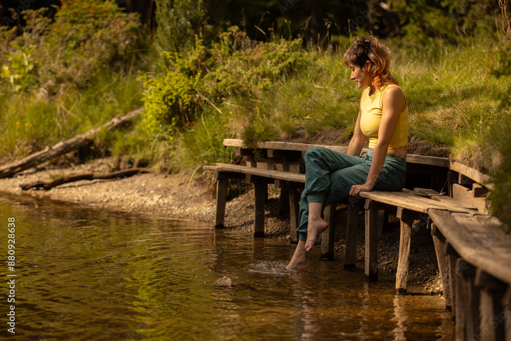 Fototapeta premium A woman sits on a bench by a body of water, her feet in the water