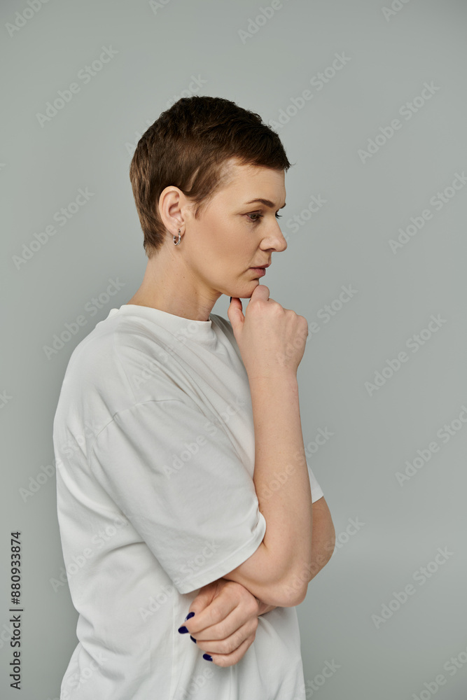 A woman with short hair, wearing casual attire and a pink ribbon, stands against a gray backdrop, thoughtfully gazing towards the side. A woman with short hair, wearing casual attire and a pink ribbon, stands against a gray backdrop, thoughtfully gazing towards the side.