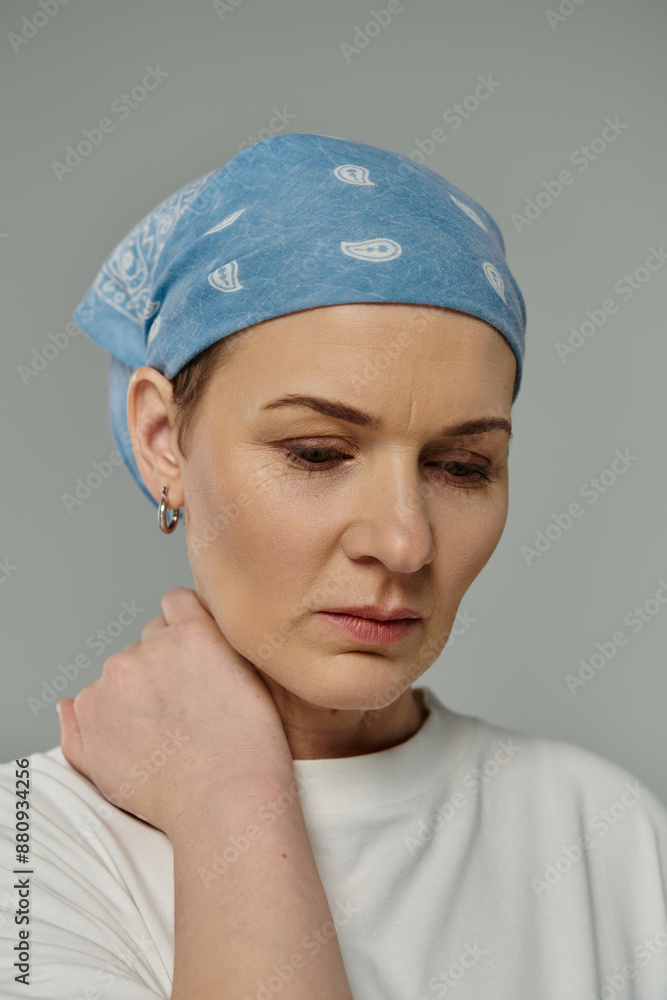 A woman with short hair and a blue bandana rests her hand on her neck, lost in thought against a grey background. A woman with short hair and a blue bandana rests her hand on her neck, lost in thought against a grey background.