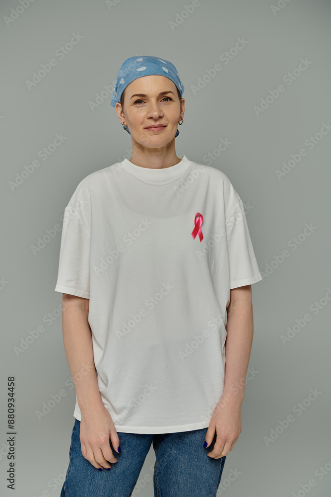 A woman with short hair, wearing a white t-shirt and blue jeans, stands against a grey background with a pink ribbon pinned to her shirt. A woman with short hair, wearing a white t-shirt and blue jeans, stands against a grey background with a pink ribbon pinned to her shirt.