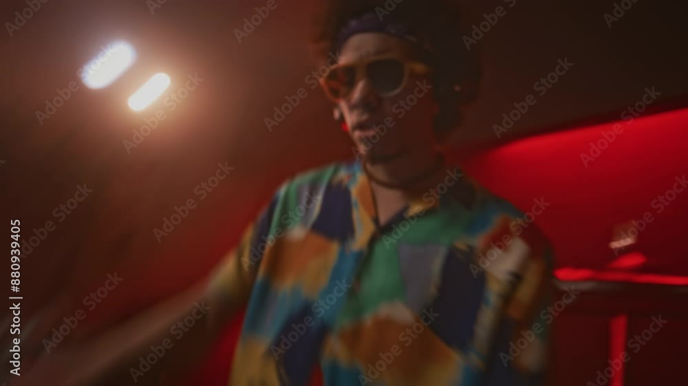 Blurred handheld shot of cool hipster guy in fancy shirt and sunglasses playing upbeat energetic music on audio mixer and dancing in red neon lit nightclub Blurred handheld shot of cool hipster guy in fancy shirt and sunglasses playing upbeat energetic music on audio mixer and dancing in red neon lit nightclub