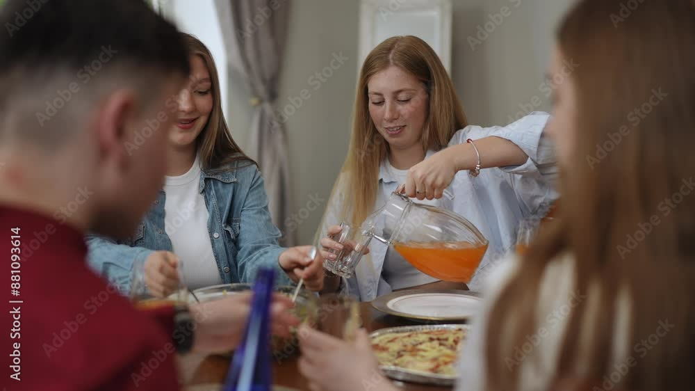 Cheerful conversation between friends at the dining table in the living room. One of the girls pours juice into a glass from a jug, and the other puts salad on her plate from a glass bowl. The company Cheerful conversation between friends at the dining table in the living room. One of the girls pours juice into a glass from a jug, and the other puts salad on her plate from a glass bowl. The company