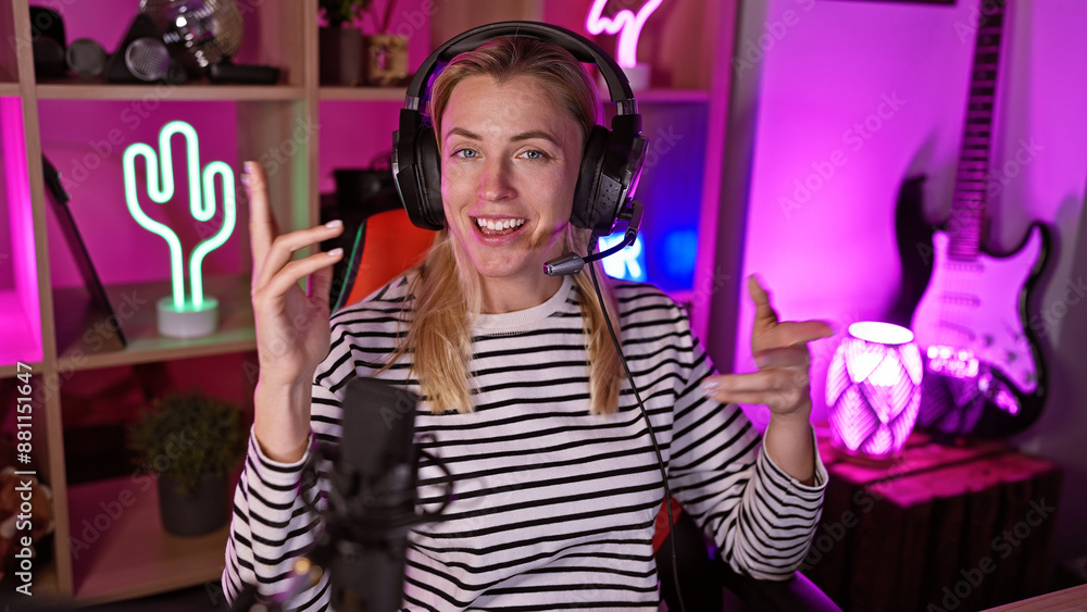 A young blonde woman wearing headphones speaks into a microphone in a vibrant gaming room at night. A young blonde woman wearing headphones speaks into a microphone in a vibrant gaming room at night.