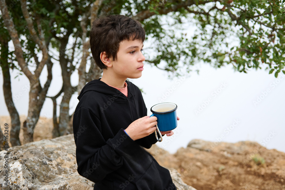 Young boy enjoying a hot drink in nature, wearing a black hoodie seated on a rock Young boy enjoying a hot drink in nature, wearing a black hoodie seated on a rock