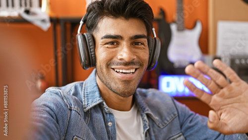 Smiling young hispanic man with beard wearing headphones in a music studio waving at the camera for a videocall
