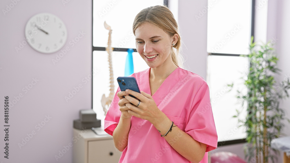 Smiling caucasian woman in pink scrubs using smartphone in bright clinic interior Smiling caucasian woman in pink scrubs using smartphone in bright clinic interior