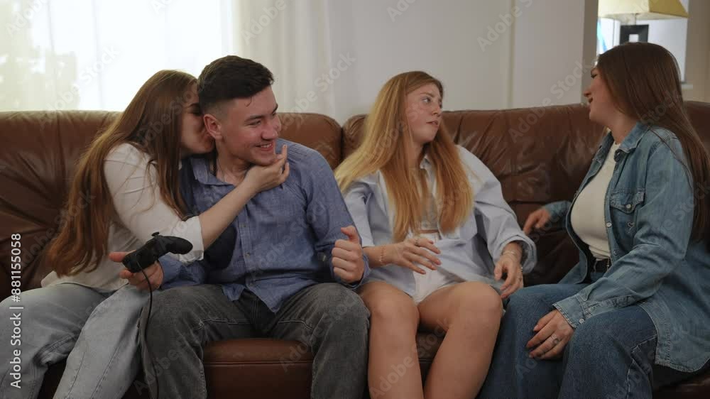 Video game sitting on a leather couch on a console in the company of friends. A joyful guy with a gamepad in his hand wins in an online game from a girl sitting next to him Video game sitting on a leather couch on a console in the company of friends. A joyful guy with a gamepad in his hand wins in an online game from a girl sitting next to him