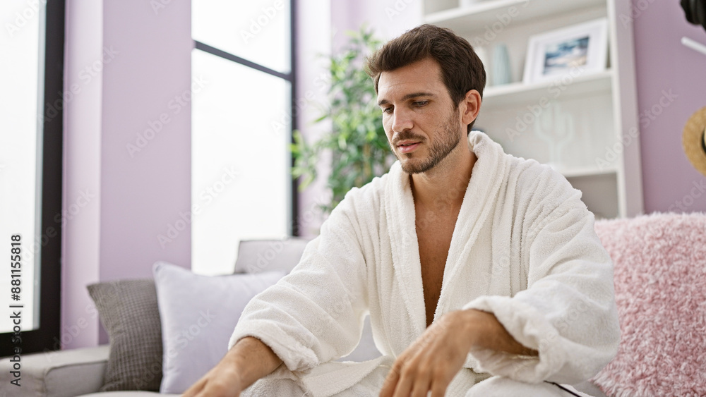 A pensive young man with a beard lounges in a robe inside a cozy modern living room, exuding casual elegance. A pensive young man with a beard lounges in a robe inside a cozy modern living room, exuding casual elegance.