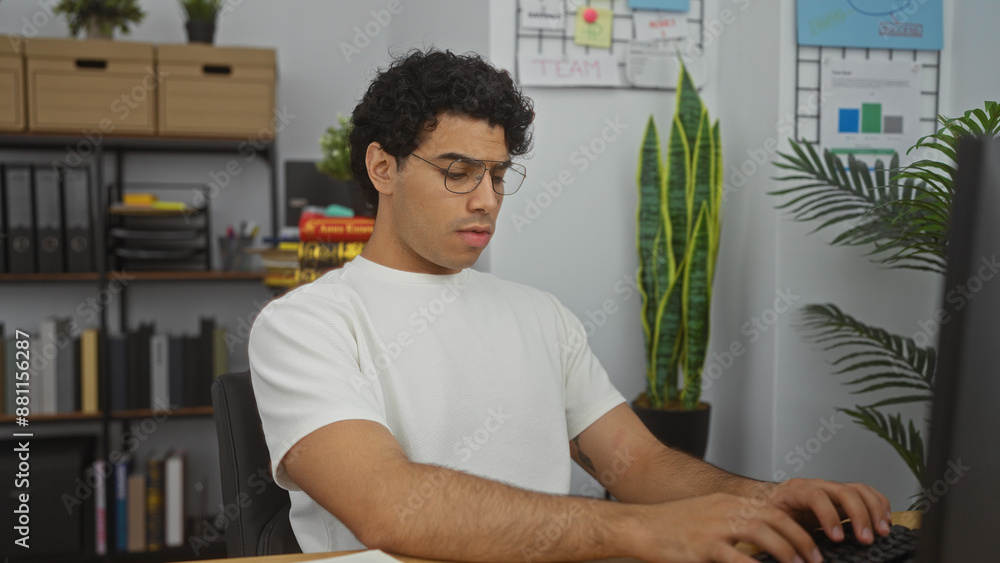 Fototapeta premium Hispanic man working in an office, focused on his computer, surrounded by organized shelves and green plants in a modern workplace.