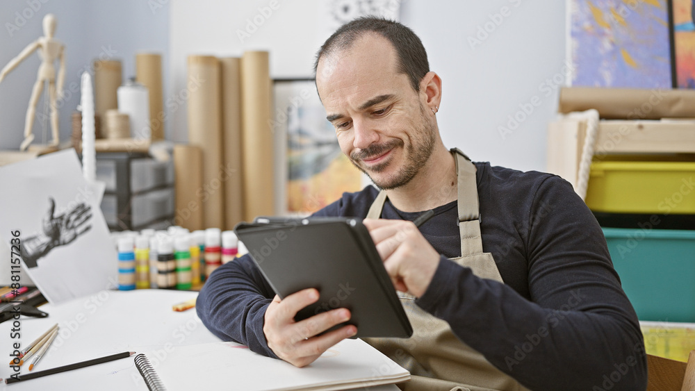 Hispanic man with beard smiles while using tablet in art studio, surrounded by painting supplies Hispanic man with beard smiles while using tablet in art studio, surrounded by painting supplies