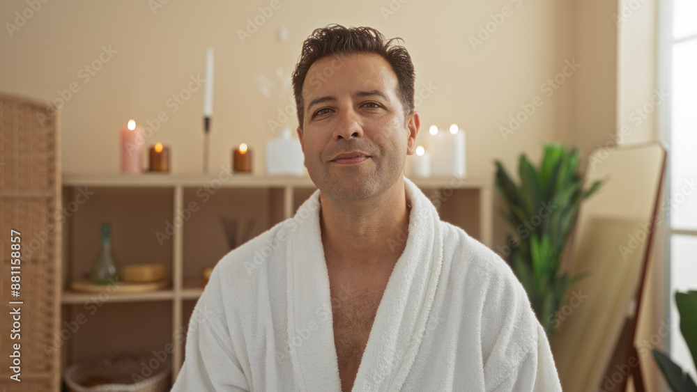 Fototapeta premium Hispanic man in a spa wearing a white robe, seated indoors with candles and plants in the background, creating a serene and relaxing ambiance in the wellness center.