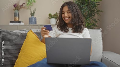 Attractive hispanic woman using laptop and holding credit card while sitting on couch in cozy living room apartment.