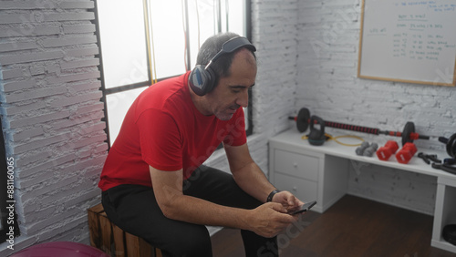 A middle-aged hispanic man in a red shirt and black pants is sitting in a gym, wearing headphones and looking at his phone.
