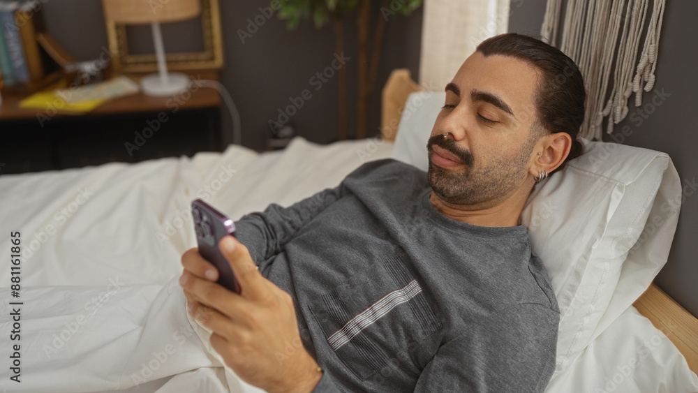 Handsome man with moustache using smartphone while lying on bed in cozy bedroom Handsome man with moustache using smartphone while lying on bed in cozy bedroom