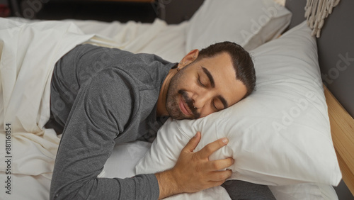 Handsome young hispanic man with a moustache sleeps comfortably in a cozy bedroom, hugging white pillows under soft blankets.