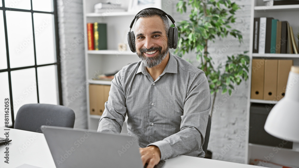 Smiling bearded senior hispanic man with headphones in an office setting using a laptop. Smiling bearded senior hispanic man with headphones in an office setting using a laptop.