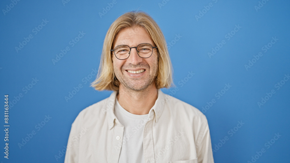 A smiling man with long blond hair and glasses posing against a blue background. A smiling man with long blond hair and glasses posing against a blue background.