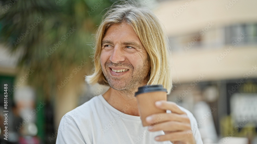 A smiling man with long blond hair holds a coffee cup while standing on a busy urban street. A smiling man with long blond hair holds a coffee cup while standing on a busy urban street.