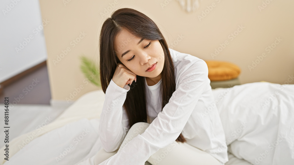 Pensive asian woman in a white shirt sitting alone in a bright bedroom reflecting on her thoughts. Pensive asian woman in a white shirt sitting alone in a bright bedroom reflecting on her thoughts.