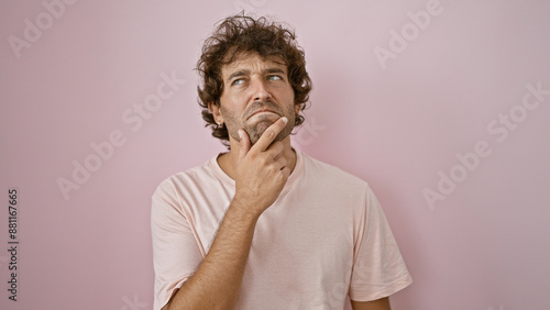 Pensive hispanic man in a casual white shirt against a plain pink background pondering with hand on chin